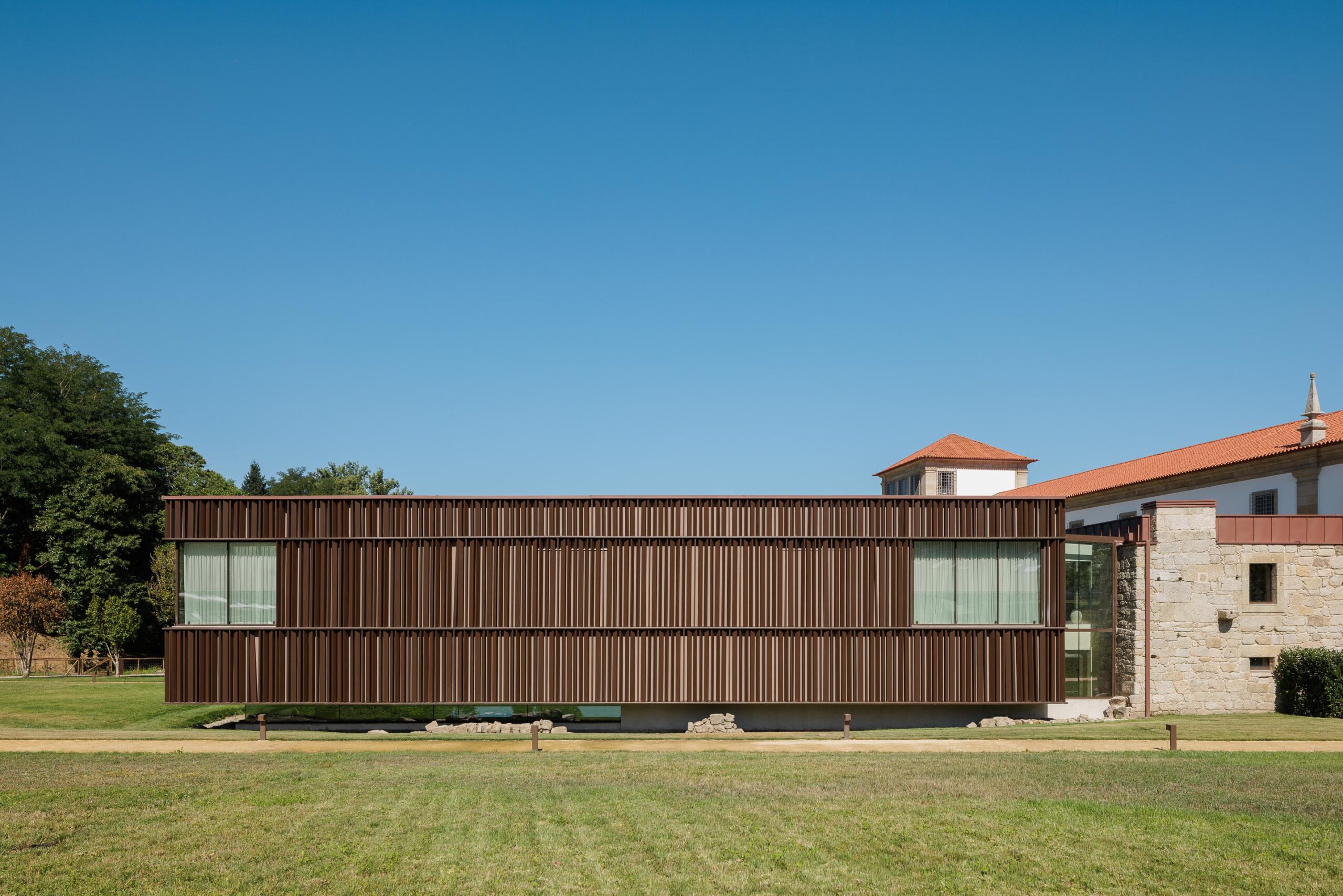 Fachada contemporânea em painéis verticais de tonalidade escura, elevada sobre embasamento transparente, junto a edifício de pedra — MS COLLECTION AROUCA arquitectura MIMOOL ARQUITECTURA & INTERIORES fotografia Ivo Tavares