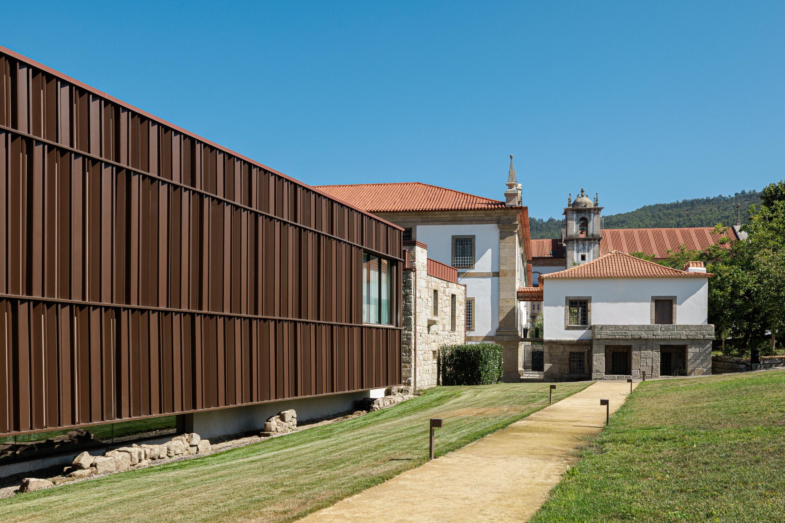 Caminho pedonal em terra batida ladeando o volume contemporâneo de fachada escura e o edifício histórico de pedra — MS COLLECTION AROUCA arquitectura MIMOOL ARQUITECTURA & INTERIORES fotografia Ivo Tavares