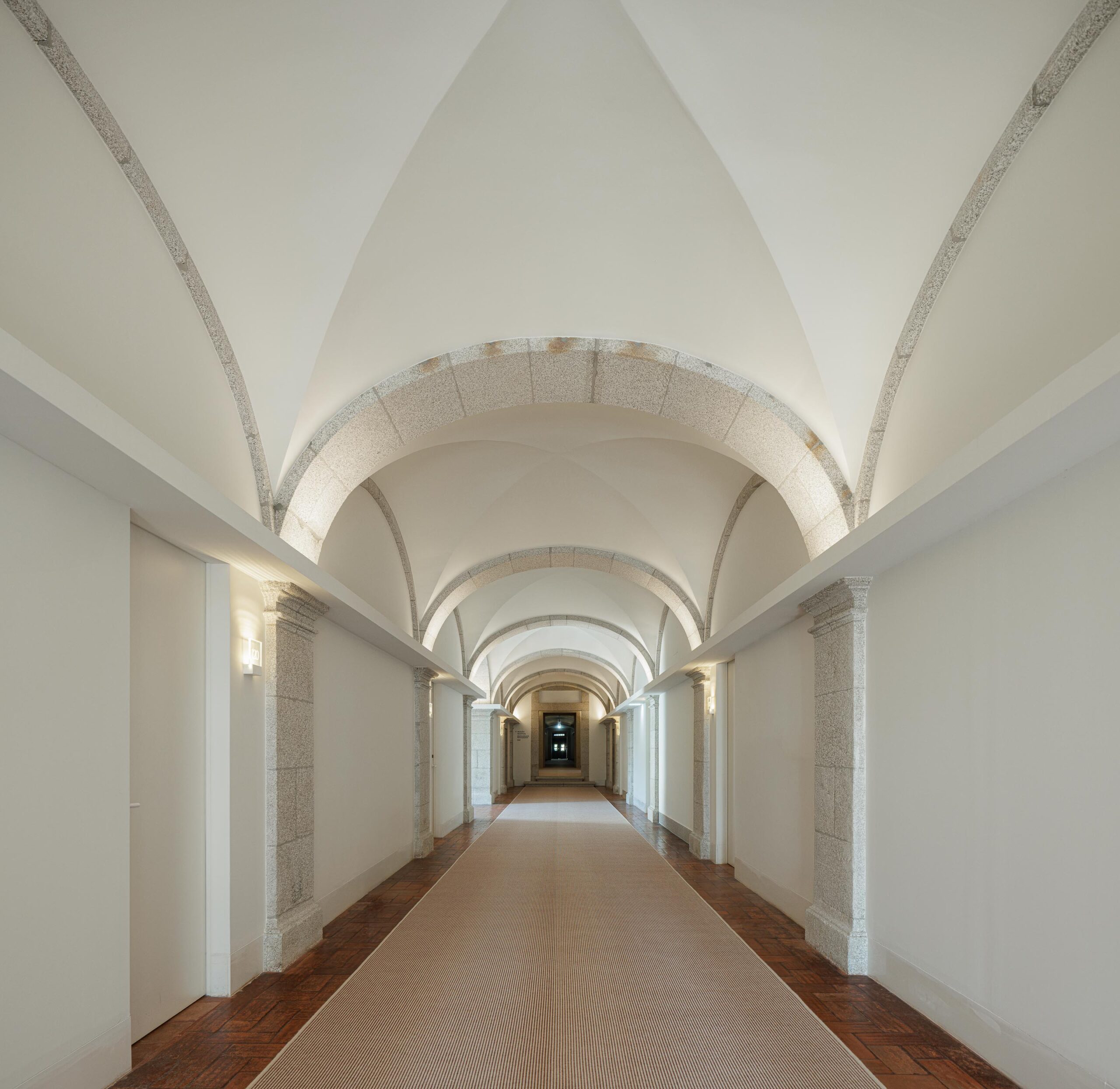 Corredor longo com abóbadas de pedra e teto branco, pavimento em madeira e tapete, criando uma perspetiva profunda — MS COLLECTION AROUCA arquitectura MIMOOL ARQUITECTURA & INTERIORES fotografia Ivo Tavares