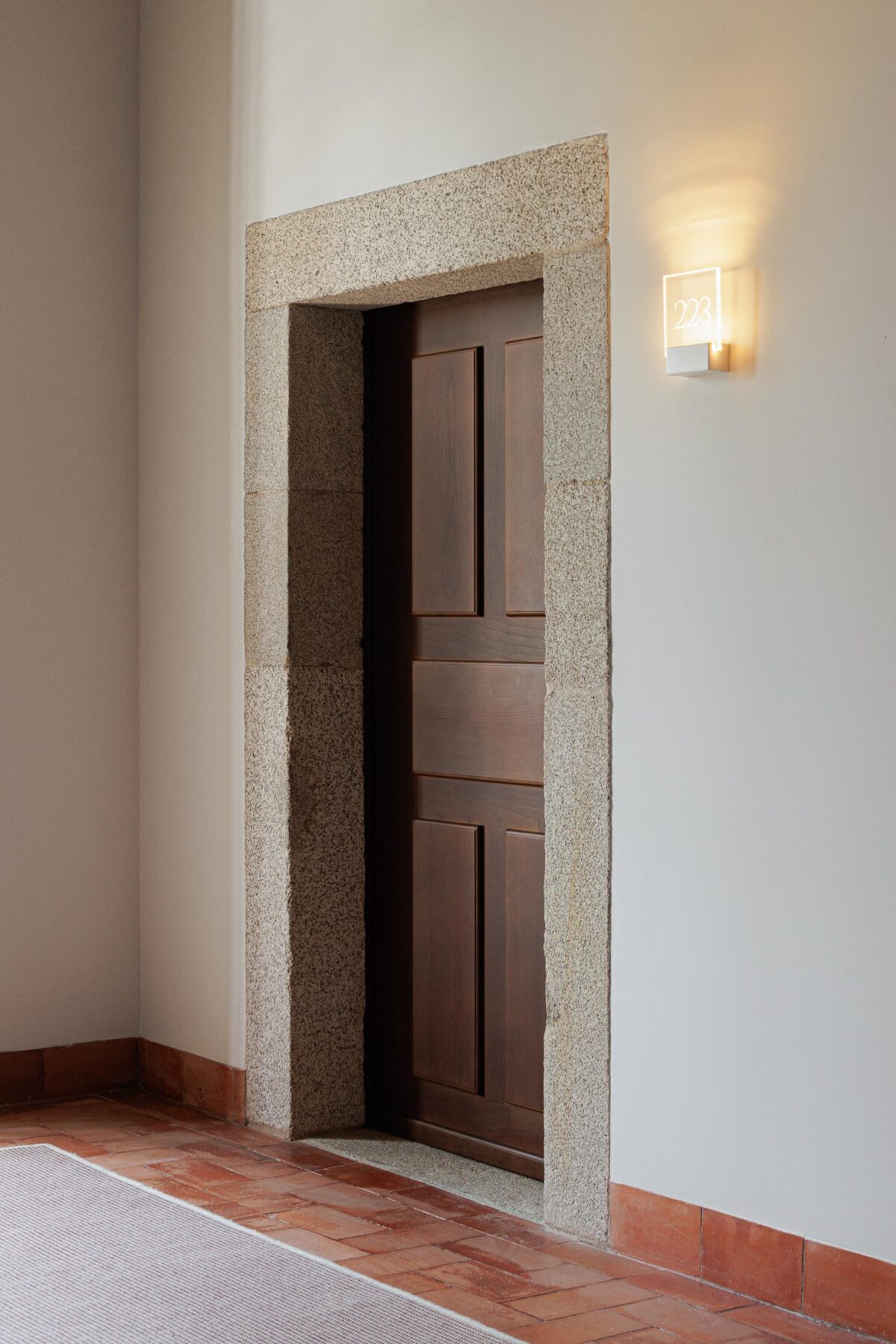 Porta em madeira escura com moldura em pedra, pavimento em tijoleira e candeeiro de parede, integrando o novo no antigo — MS COLLECTION AROUCA arquitectura MIMOOL ARQUITECTURA & INTERIORES fotografia Ivo Tavares