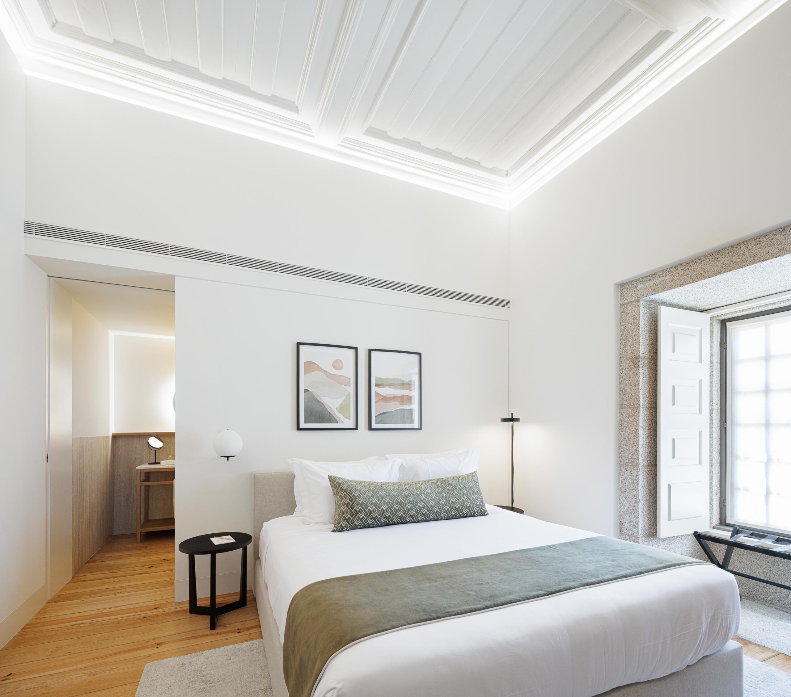Quarto com cama, pavimento em madeira clara, teto branco trabalhado e janela em pedra, com acesso à casa de banho — MS COLLECTION AROUCA arquitectura MIMOOL ARQUITECTURA & INTERIORES fotografia Ivo Tavares