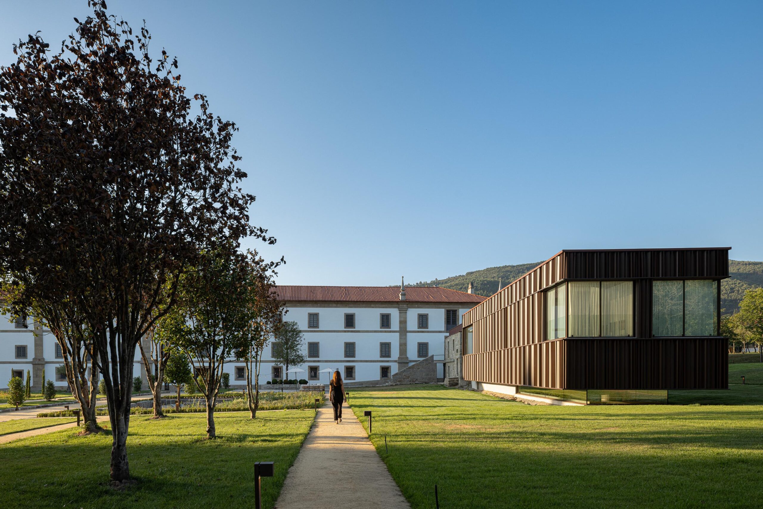 Caminho pedonal em relva, ladeado pelo edifício histórico e o novo volume em lamelas metálicas — MS COLLECTION AROUCA arquitectura MIMOOL ARQUITECTURA & INTERIORES fotografia Ivo Tavares