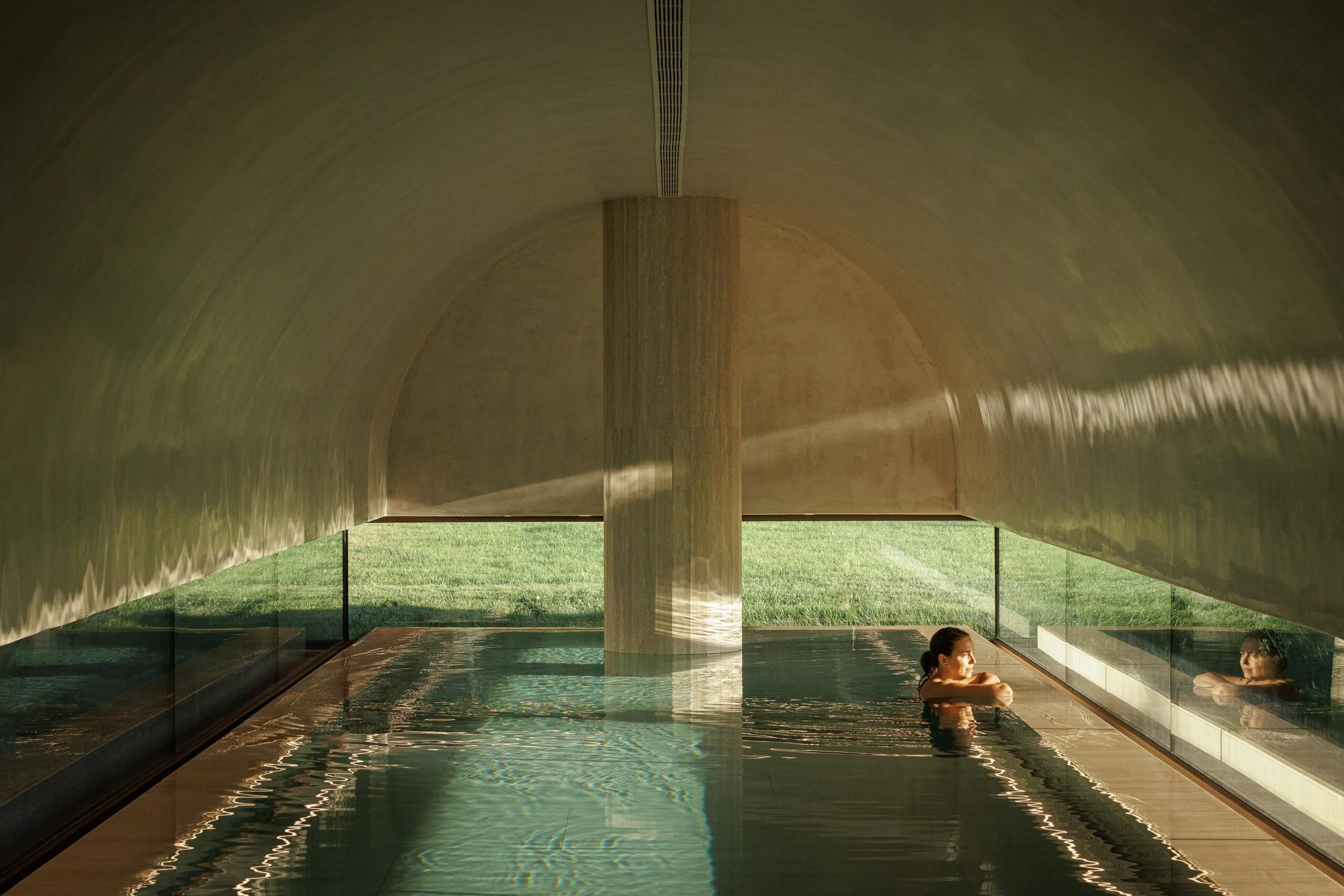 Piscina interior com teto abobadado, coluna central e vista panorâmica para a relva, com luz natural — MS COLLECTION AROUCA arquitectura MIMOOL ARQUITECTURA & INTERIORES fotografia Ivo Tavares