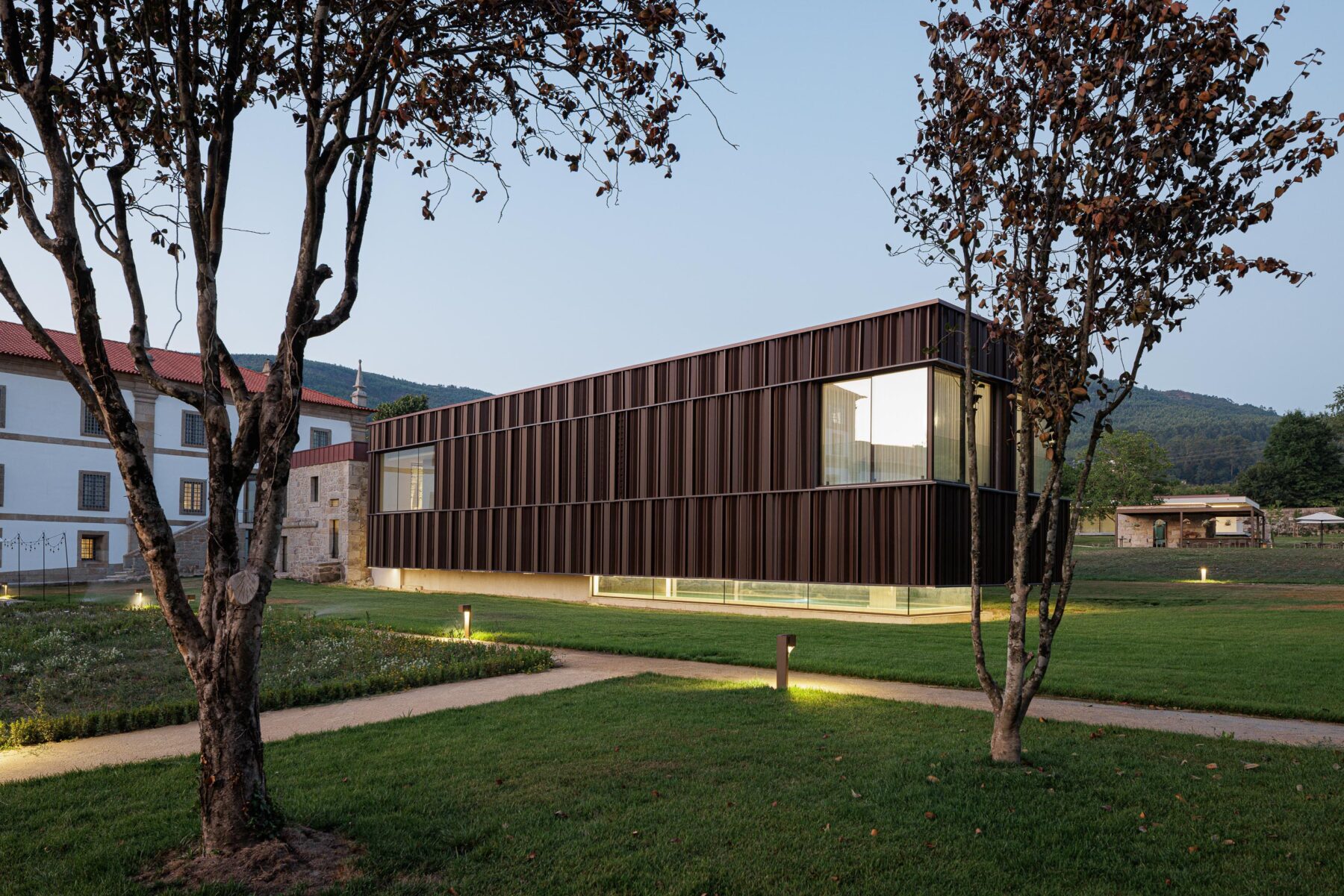 Vista noturna do novo volume em lamelas metálicas e vidro, iluminado, com o edifício histórico ao fundo — MS COLLECTION AROUCA arquitectura MIMOOL ARQUITECTURA & INTERIORES fotografia Ivo Tavares