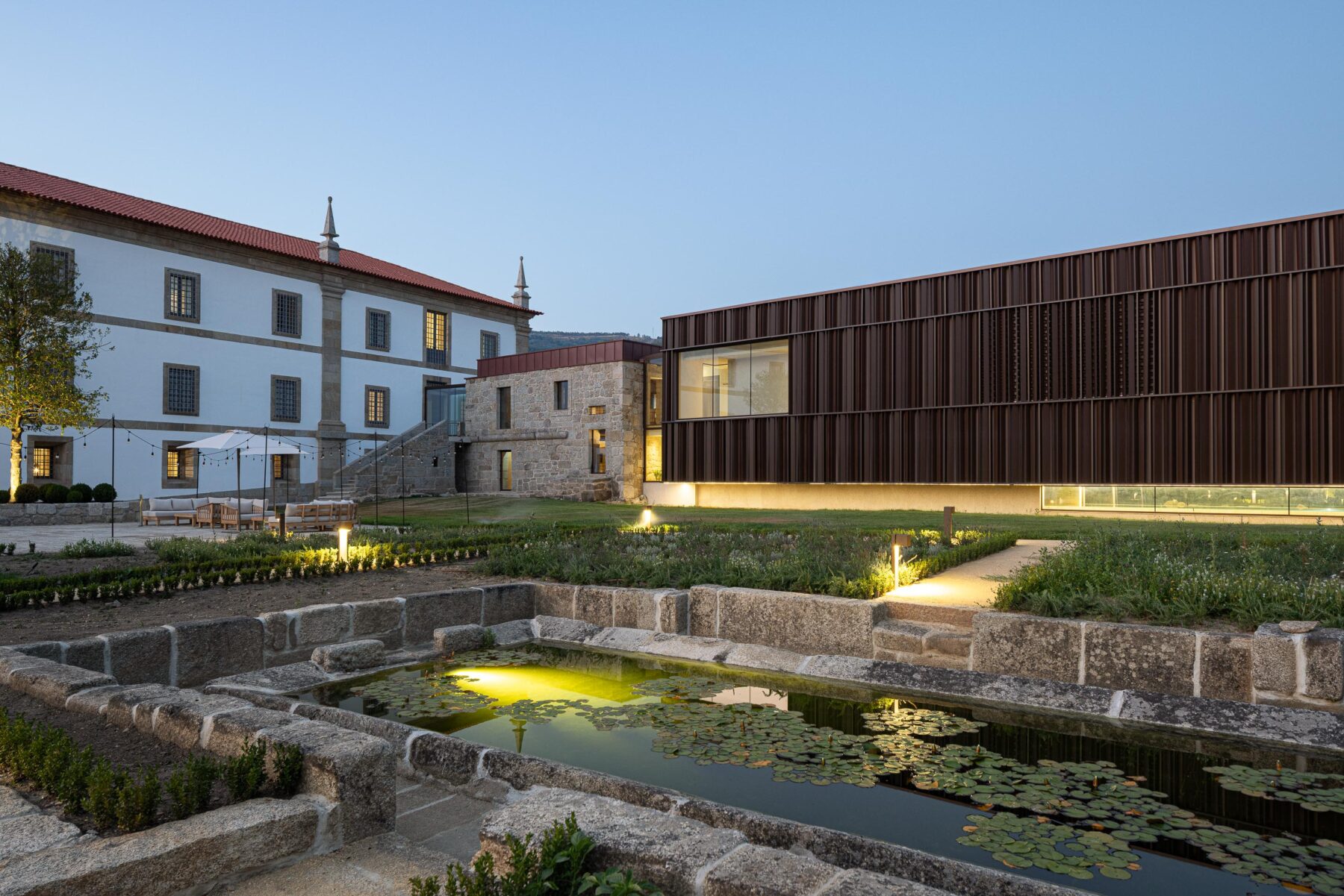 Vista noturna do tanque em pedra e do novo volume em lamelas metálicas, com o edifício histórico ao fundo — MS COLLECTION AROUCA arquitectura MIMOOL ARQUITECTURA & INTERIORES fotografia Ivo Tavares