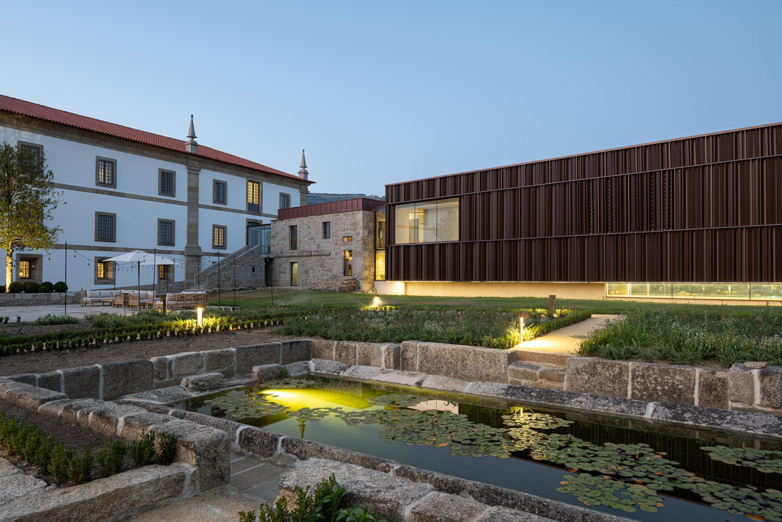 Vista noturna do tanque em pedra e do novo volume em lamelas metálicas, com o edifício histórico ao fundo — MS COLLECTION AROUCA arquitectura MIMOOL ARQUITECTURA & INTERIORES fotografia Ivo Tavares