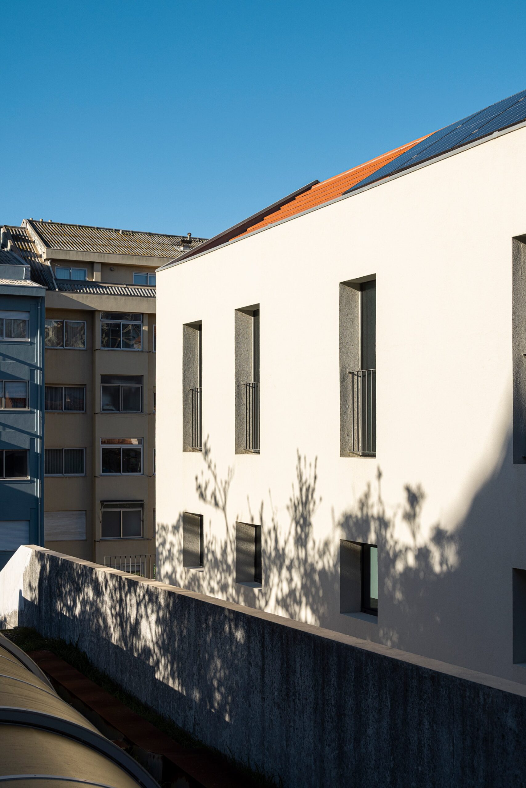 Fachada lateral da moradia com revestimento branco, vãos verticais e sombras projetadas de vegetação — Casa sem Rua