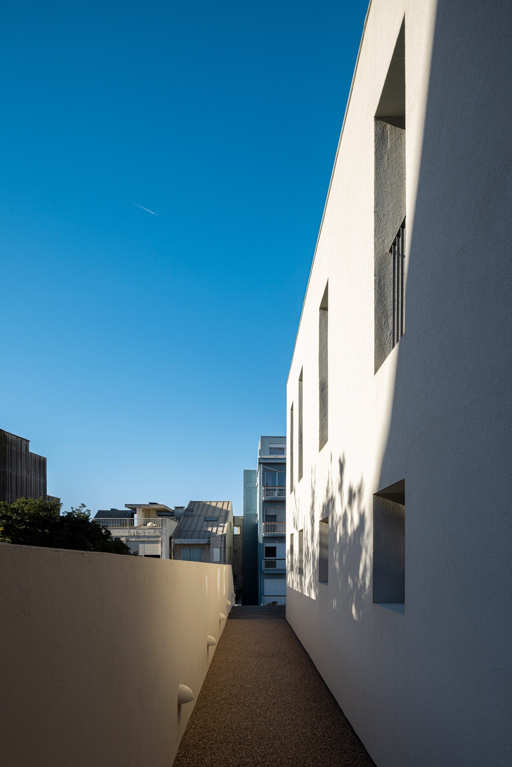 Corredor exterior estreito com pavimento em gravilha e paredes brancas, ladeado por edifícios — Casa sem Rua