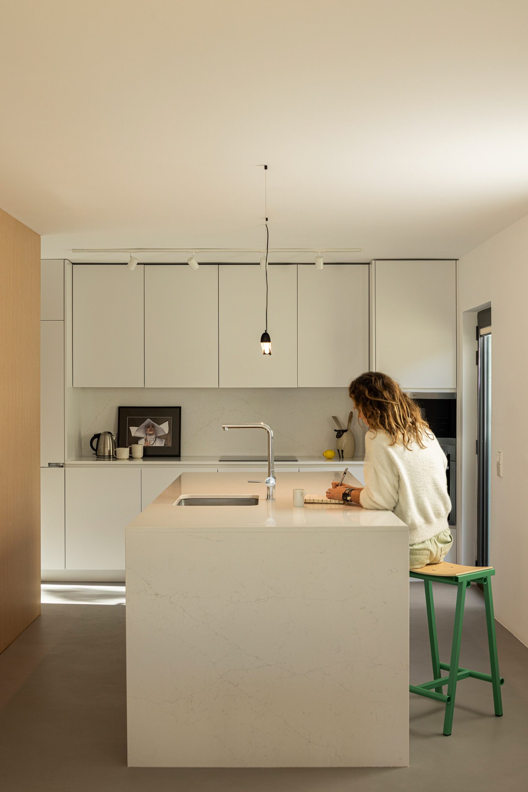 Cozinha com ilha central em mármore branco, armários brancos e pavimento cinzento, com luz natural — Casa sem Rua