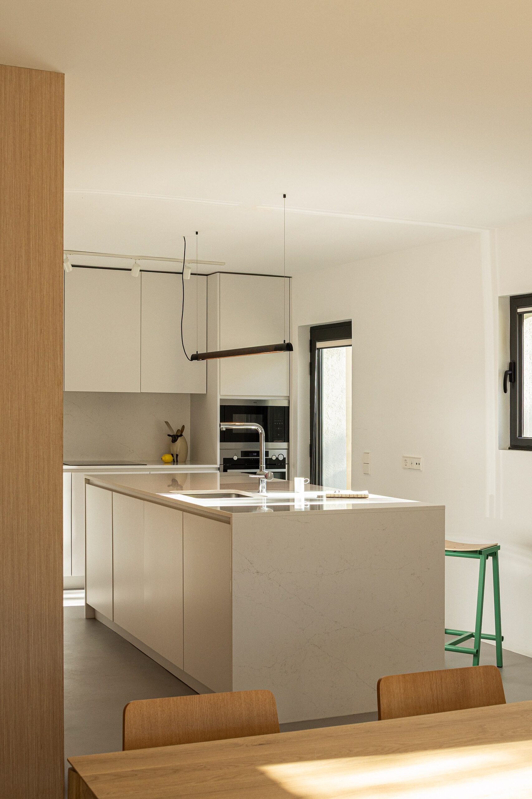 Cozinha com ilha central em mármore branco, armários brancos e pavimento cinzento, com luz natural — Casa sem Rua