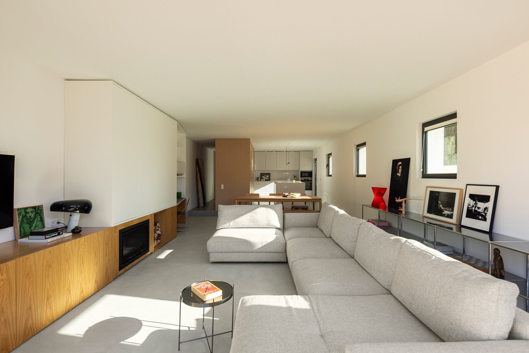 Sala de estar ampla com sofá cinzento, pavimento cinzento e armários em madeira clara e branco — Casa sem Rua