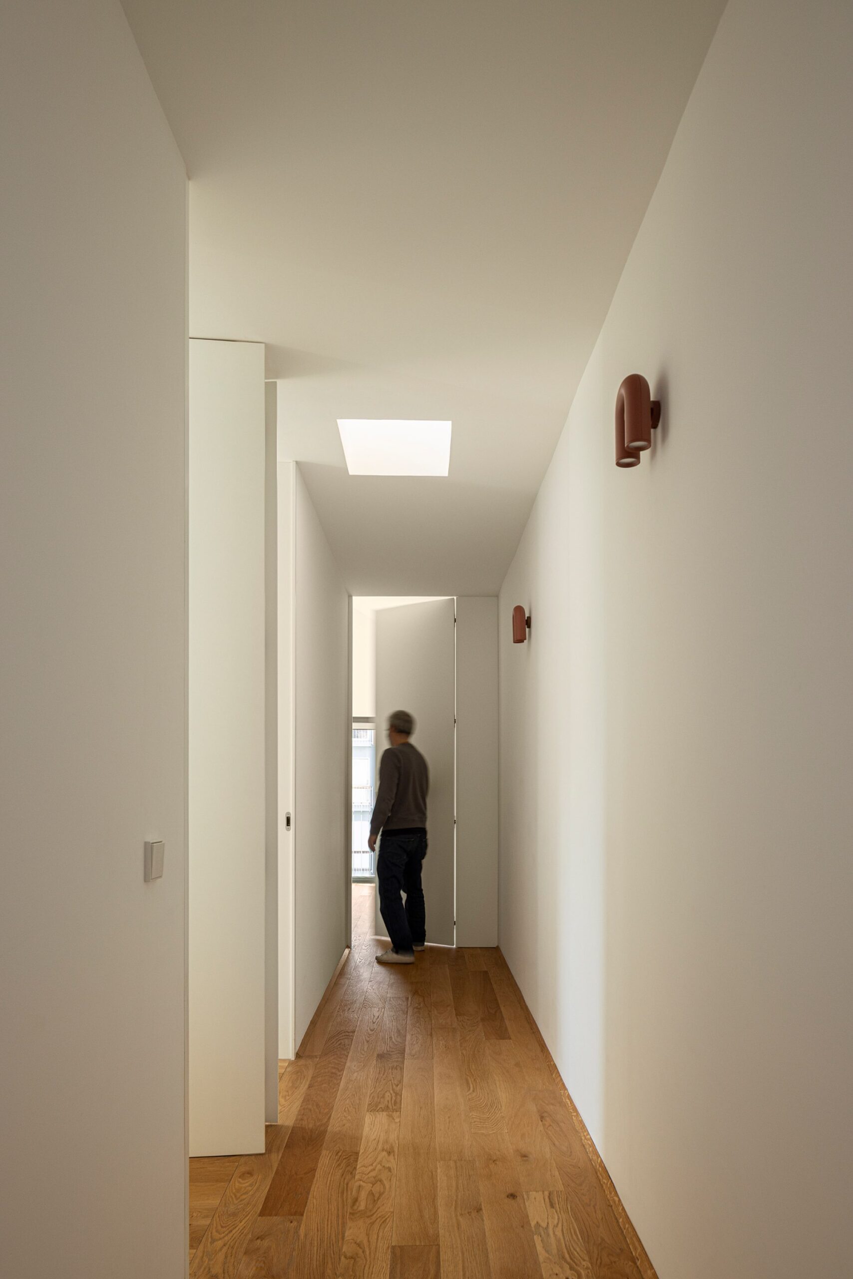 Corredor interior com pavimento em madeira de carvalho, iluminação zenital e candeeiros de parede — Casa sem Rua