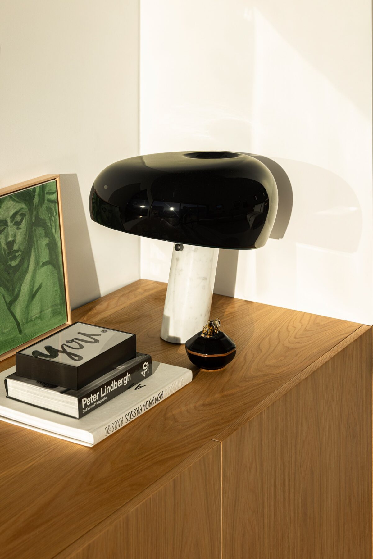 Detalhe de candeeiro preto sobre móvel em madeira clara, com livros e quadro — Casa sem Rua