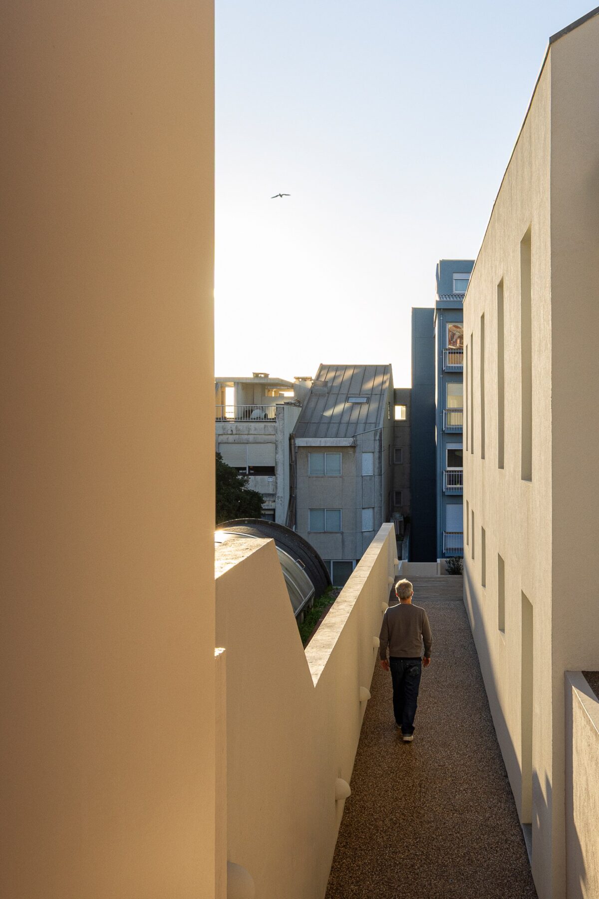 Corredor exterior com pavimento em gravilha, paredes brancas e luz do sol — Casa sem Rua