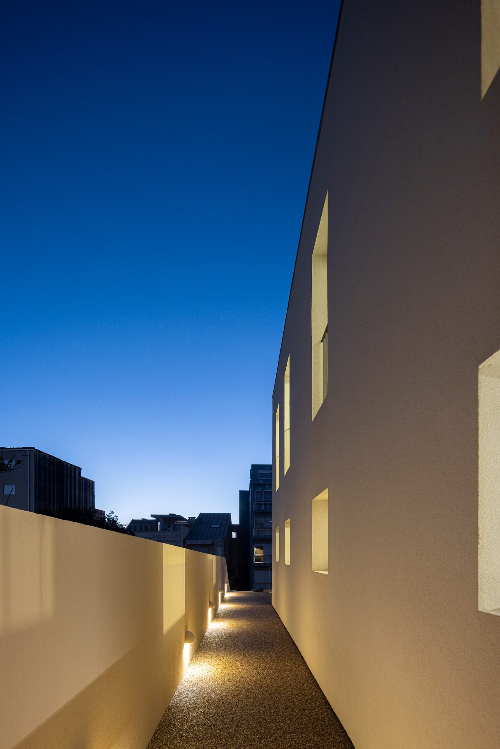 Corredor exterior com pavimento em gravilha, paredes brancas e iluminação embutida — Casa sem Rua