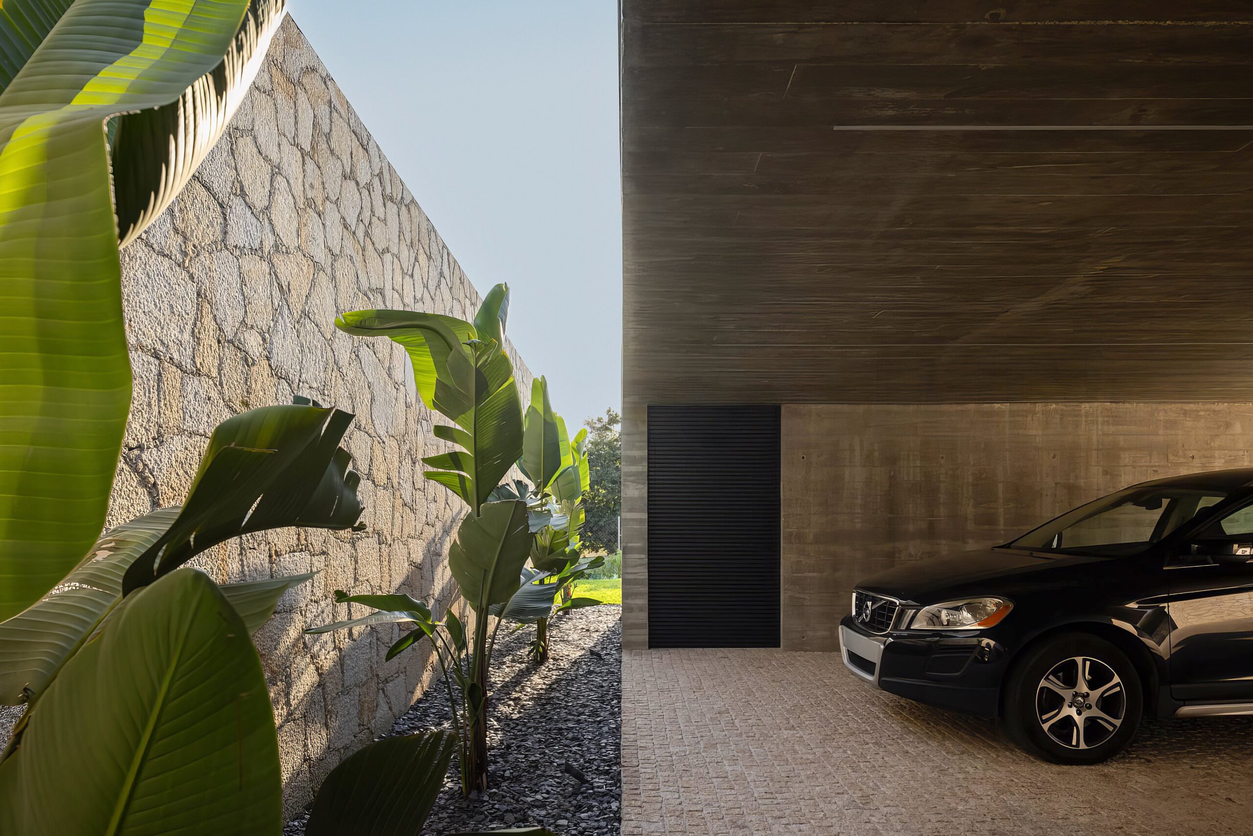 Garagem com cobertura em betão aparente, pavimento em calçada e parede em alvenaria de granito, com vegetação — Quinta da Casa (Habitação QC)