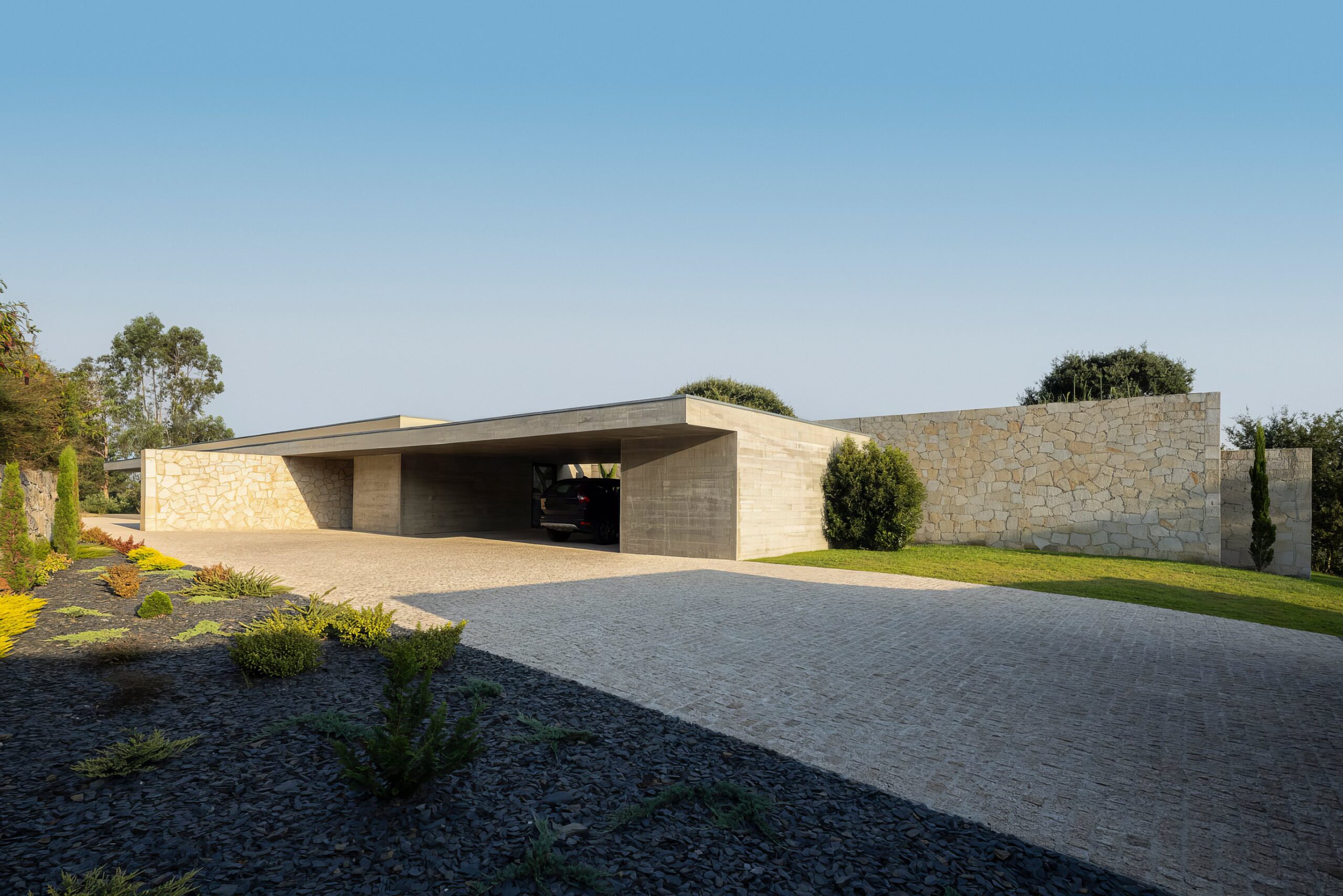 Acesso à moradia com pavimento em calçada, volumes em betão aparente e alvenaria de granito, e vegetação — Quinta da Casa (Habitação QC)