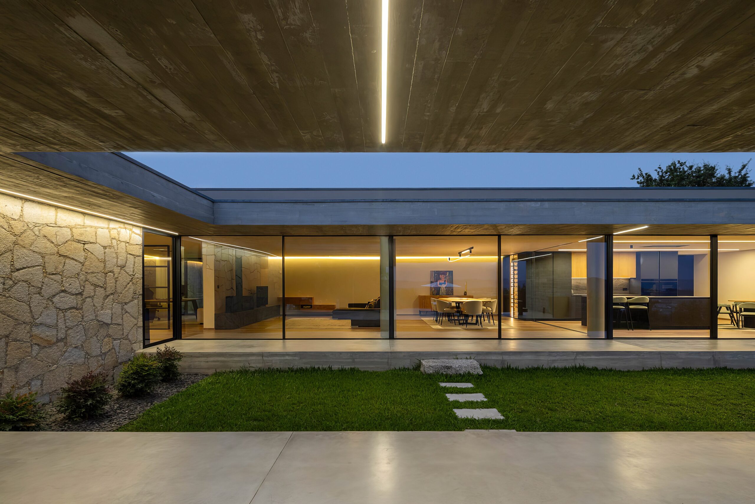 Pátio interior da moradia ao entardecer, com pavimento em relva, paredes em alvenaria de granito e iluminação linear — Quinta da Casa (Habitação QC)