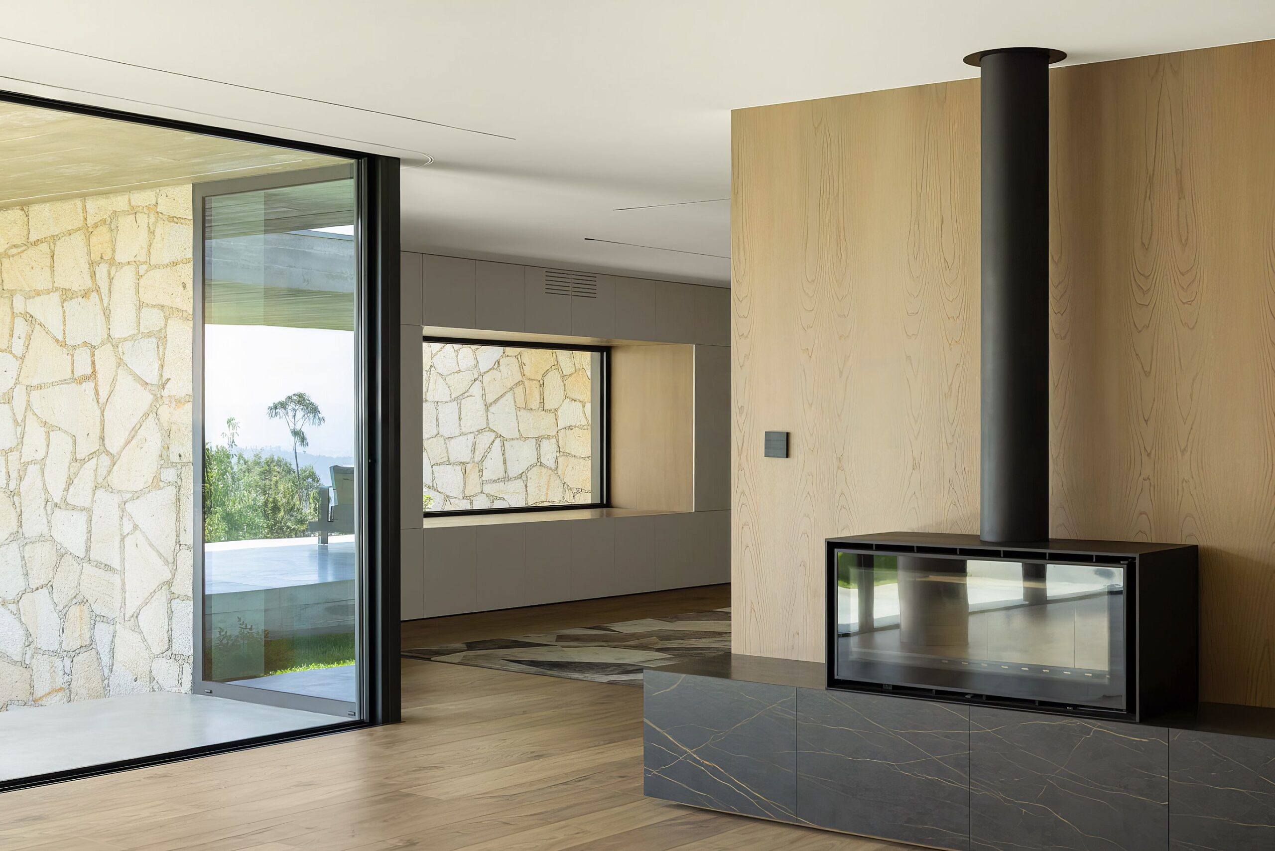 Sala com lareira em mármore negro e madeira mel, pavimento em madeira e vãos envidraçados para o exterior — Quinta da Casa (Habitação QC)