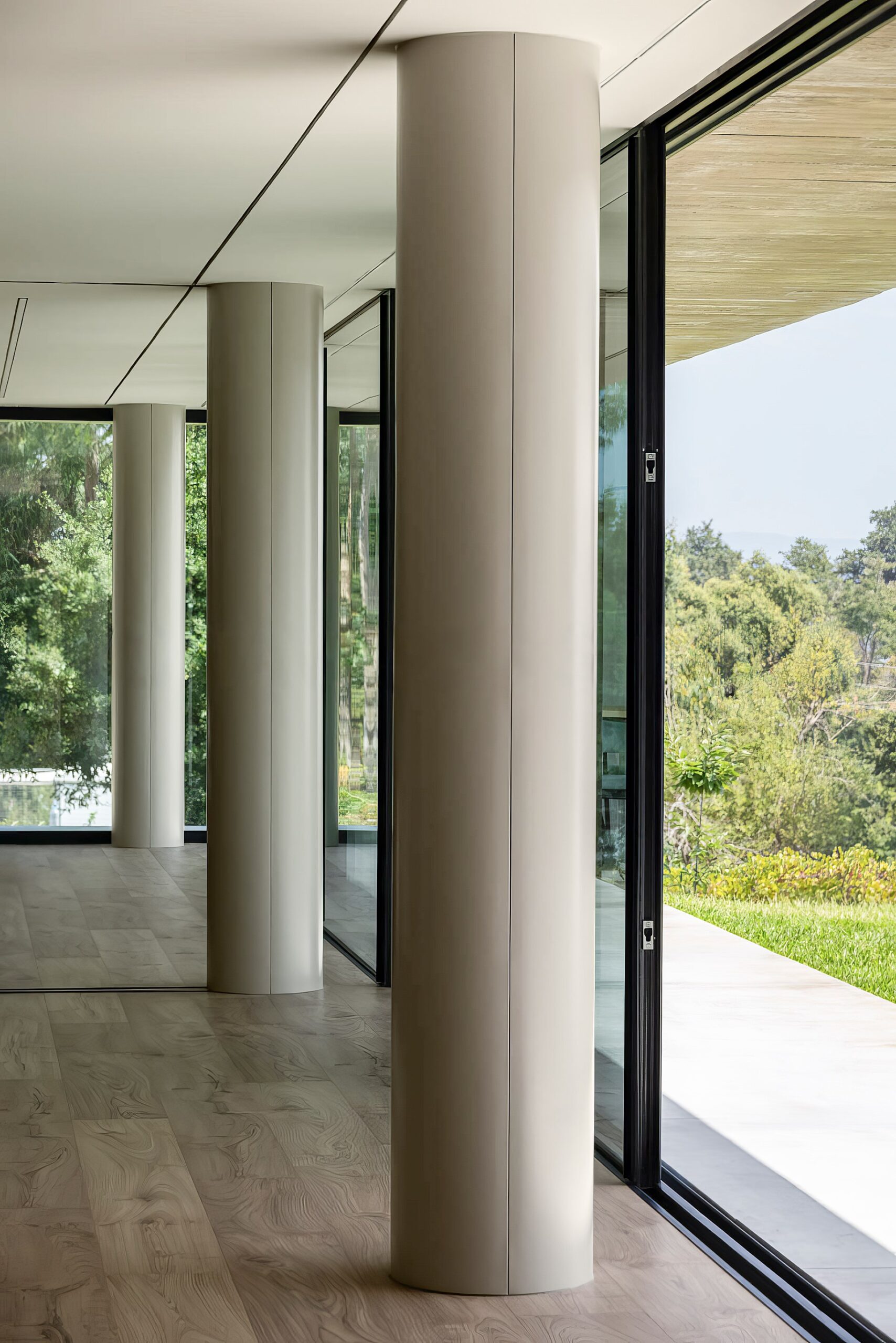 Interior da moradia com pavimento em madeira, pilares cilíndricos e grandes vãos envidraçados para o exterior — Quinta da Casa (Habitação QC)