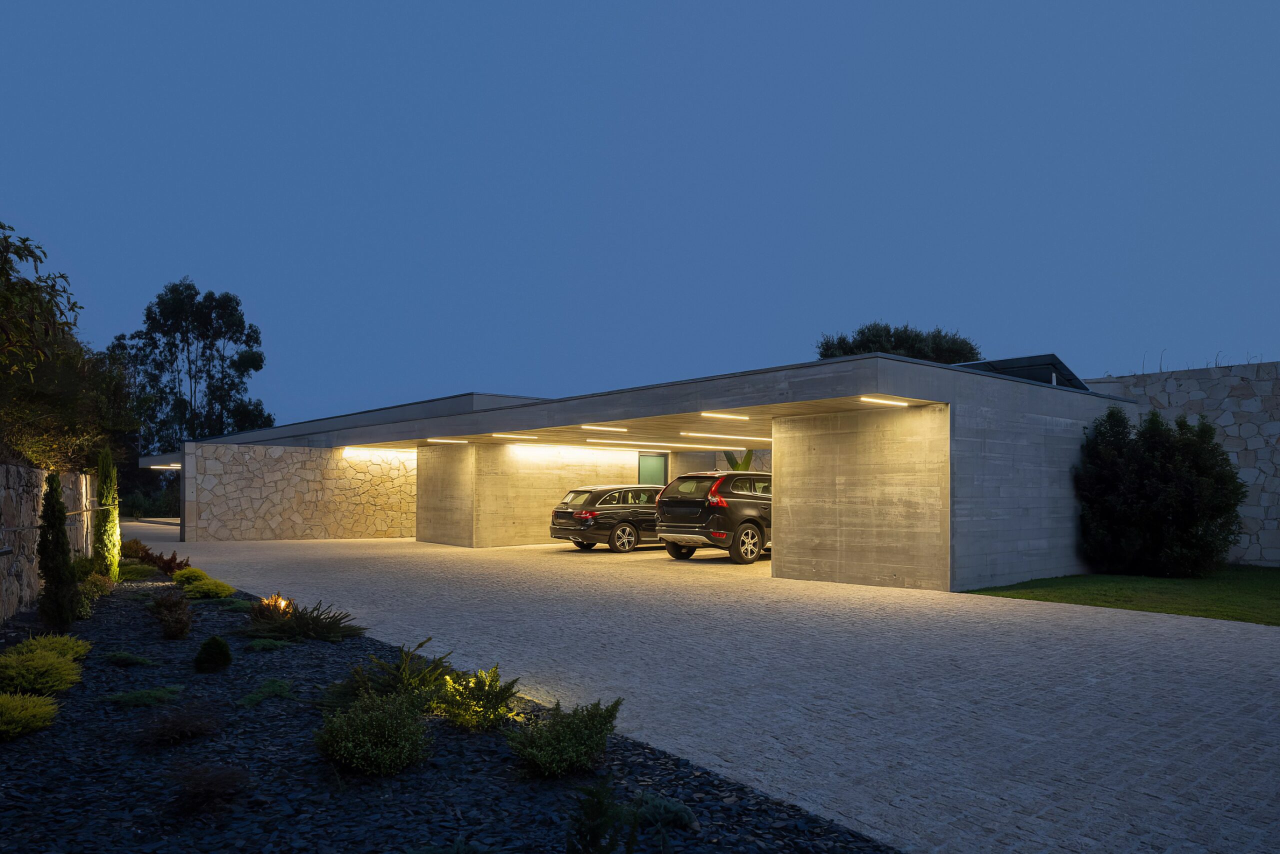 Entrada da moradia ao entardecer, com alpendre em betão aparente, pavimento em calçada e iluminação linear — Quinta da Casa (Habitação QC)