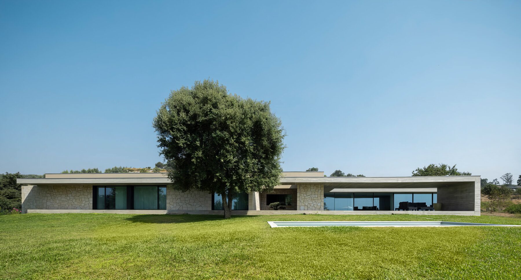 Fachada de moradia térrea em betão aparente e alvenaria de granito, com grandes vãos envidraçados e piscina — Quinta da Casa (Habitação QC)