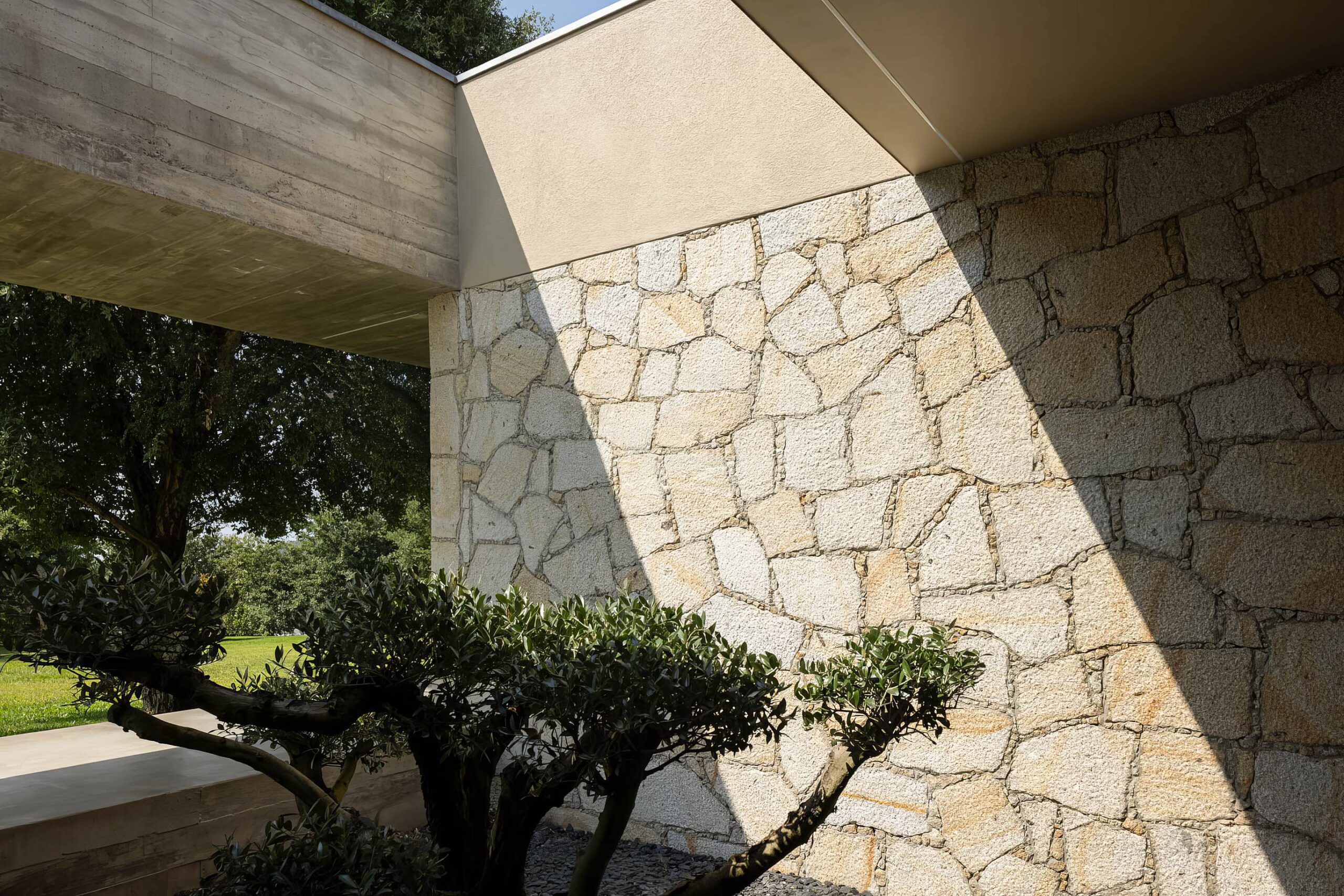 Detalhe da fachada em alvenaria de granito com betão aparente e vegetação, sob luz solar — Quinta da Casa (Habitação QC)
