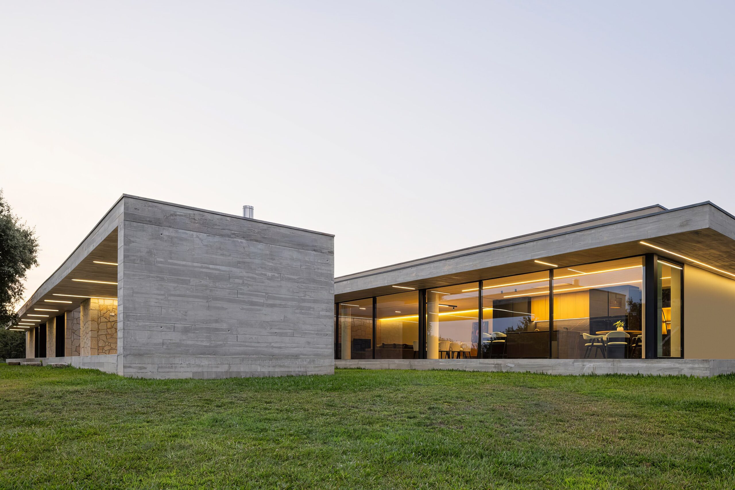 Fachada da moradia ao entardecer, com volumes em betão aparente e alvenaria de granito, e iluminação interior — Quinta da Casa (Habitação QC)