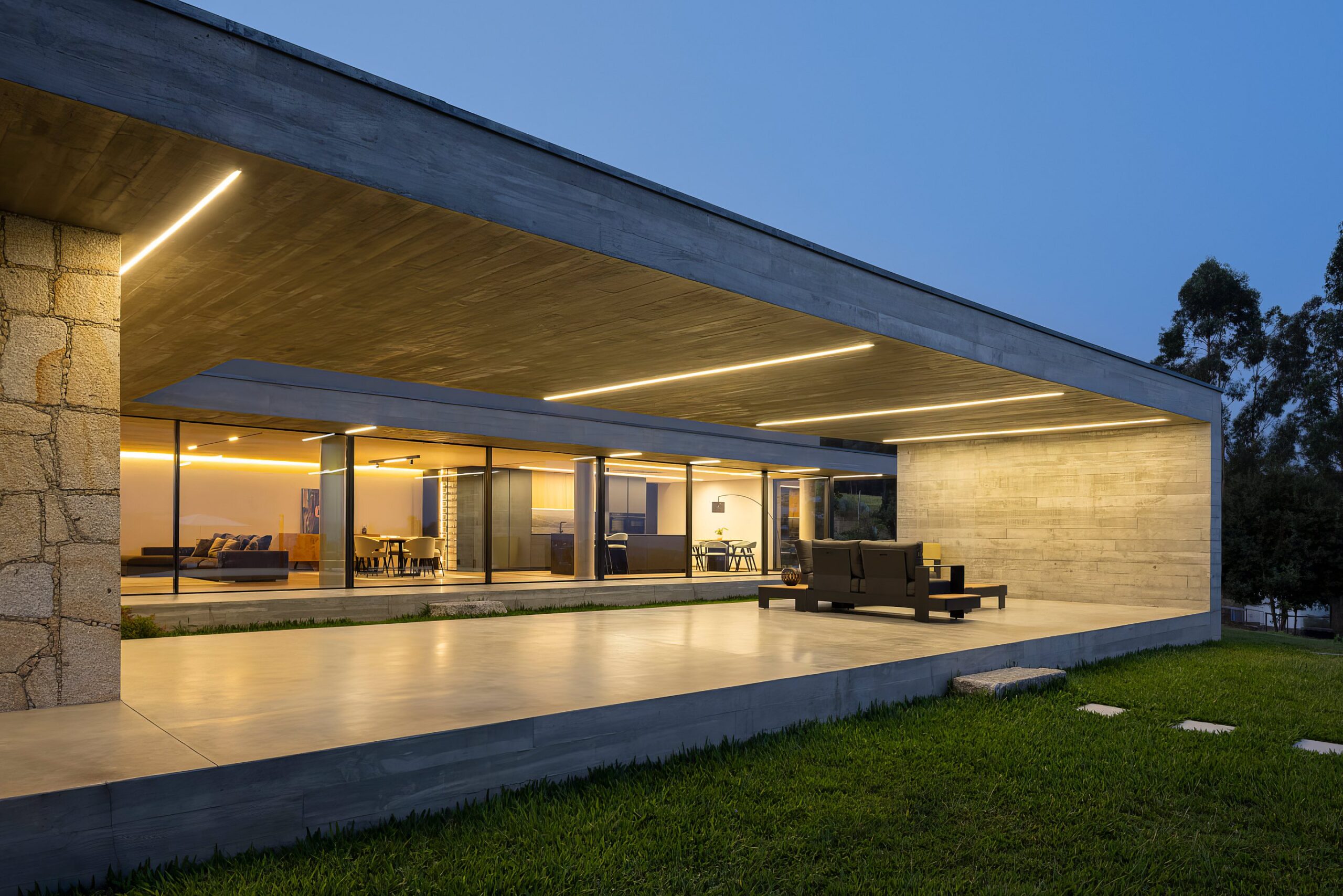 Alpendre exterior da moradia ao entardecer, com pavimento em betão aparente, mobiliário e iluminação linear — Quinta da Casa (Habitação QC)