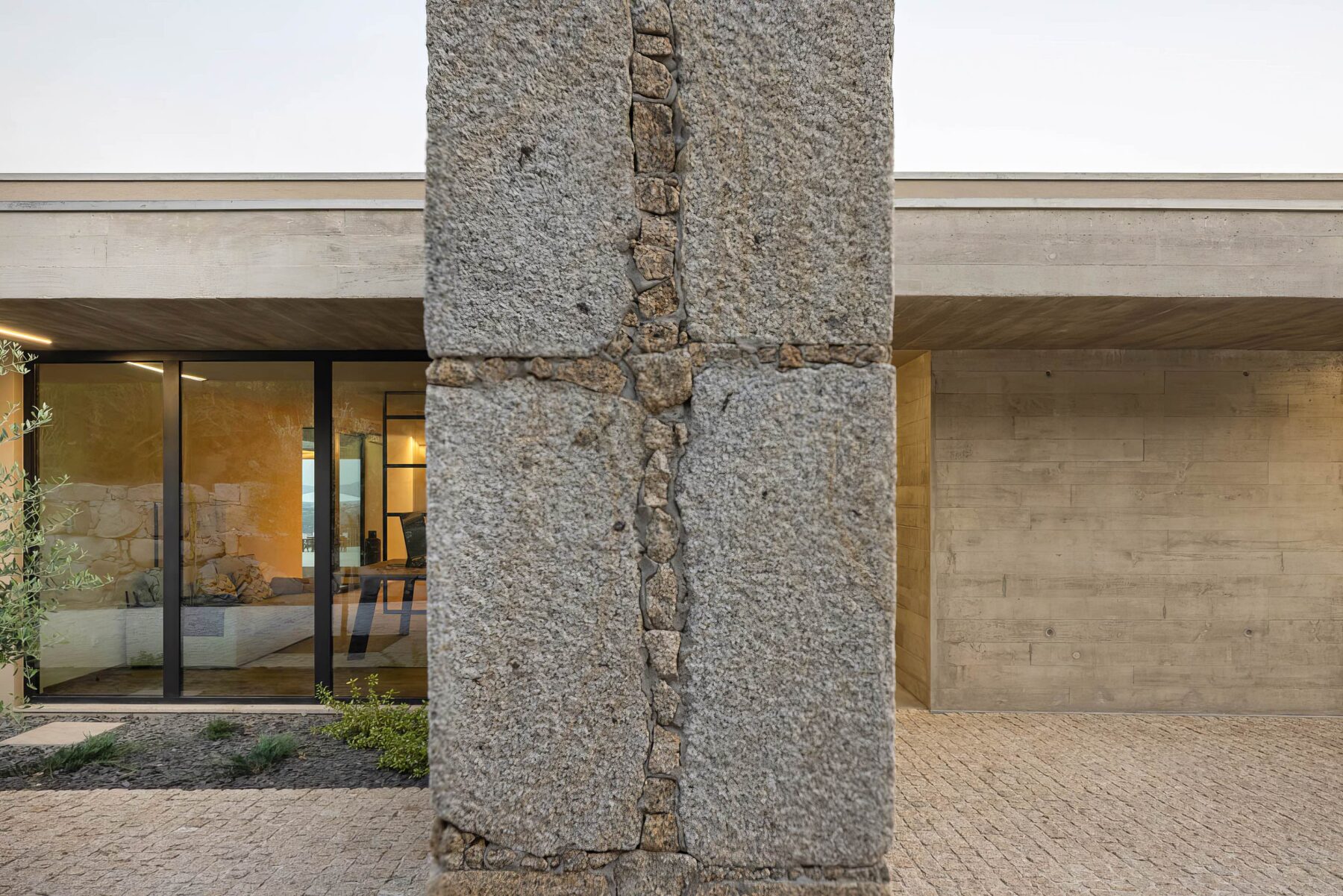 Detalhe de pilar em granito rústico, com betão aparente e pavimento em calçada, na entrada da moradia — Quinta da Casa (Habitação QC)