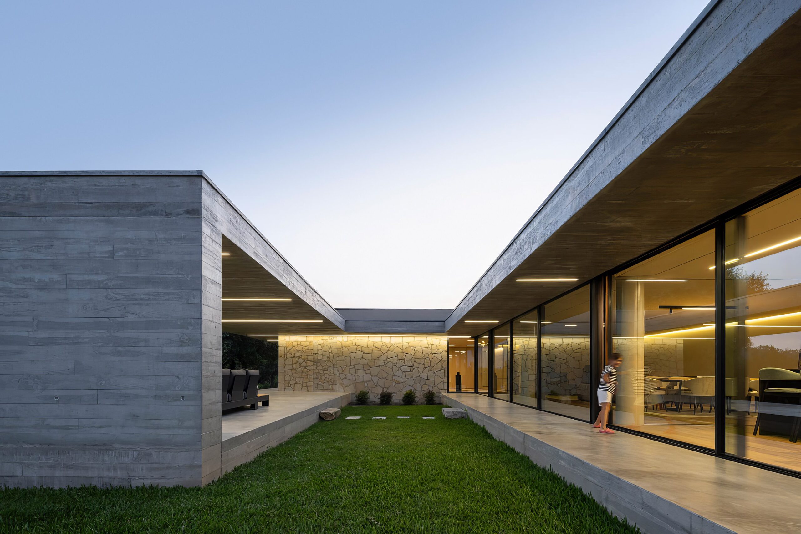 Pátio interior da moradia com pavimento em relva, paredes em alvenaria de granito e betão aparente, e iluminação linear — Quinta da Casa (Habitação QC)