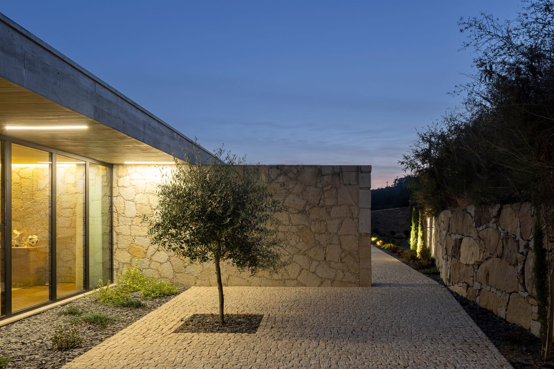 Caminho de acesso à moradia ao entardecer, com pavimento em calçada, paredes em alvenaria de granito e iluminação — Quinta da Casa (Habitação QC)