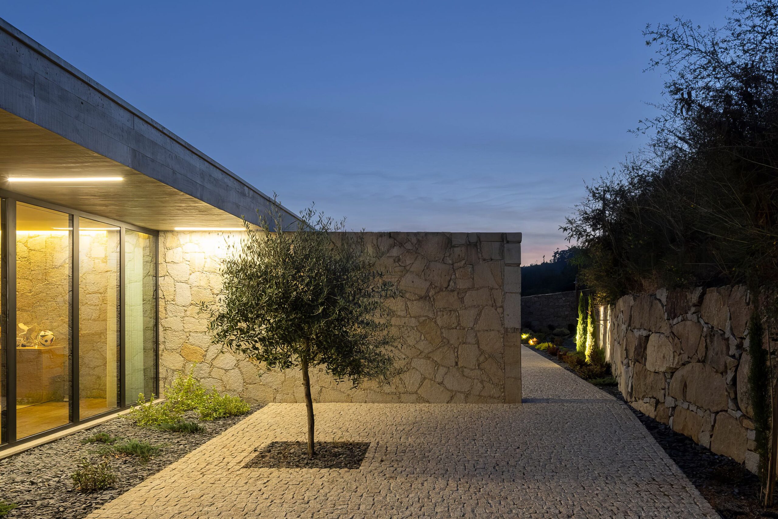 Caminho de acesso à moradia ao entardecer, com pavimento em calçada, paredes em alvenaria de granito e iluminação — Quinta da Casa (Habitação QC)